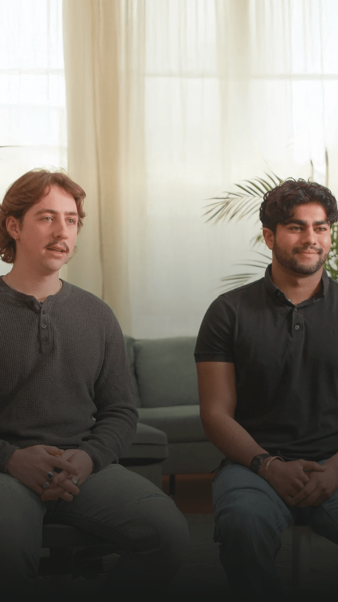 Two people seated side by side in a sunlit room with sheer curtains and a houseplant behind them.