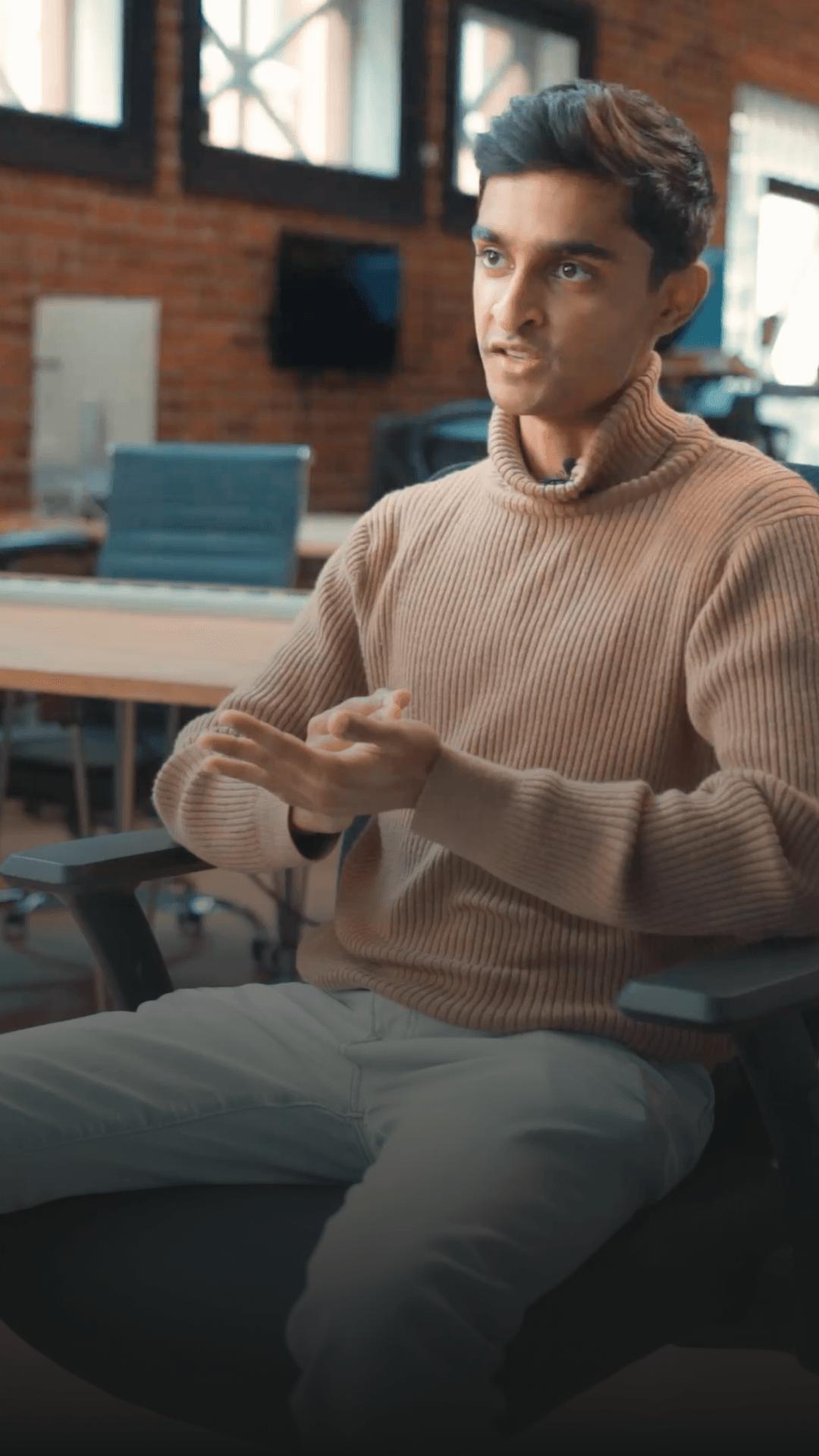 A person in a tan turtleneck sweater speaking while seated in an office chair in a brick-walled workspace.