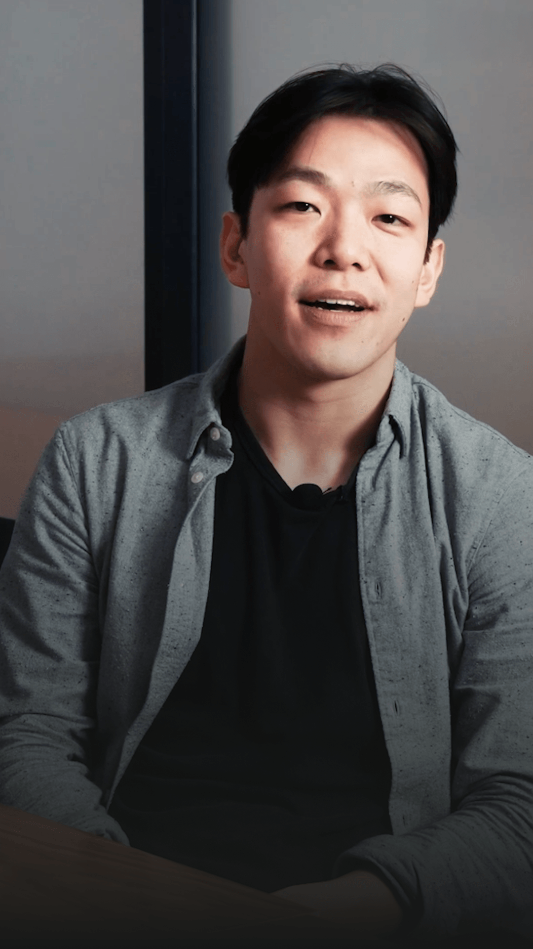 A person in a grey overshirt speaking while seated beside a window.