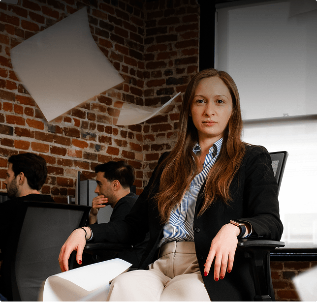 A person in a dark blazer seated in a brick-walled office, with loose papers suspended in the air behind them and two colleagues working in the background.
