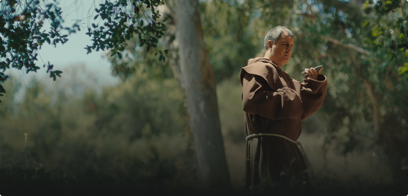 A man in a brown robe stands outdoors, looking at a smartphone, surrounded by trees and greenery.