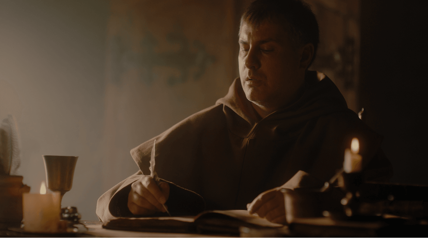 A monk in a dimly lit room writes in a book by candlelight, surrounded by candles and a metal cup.