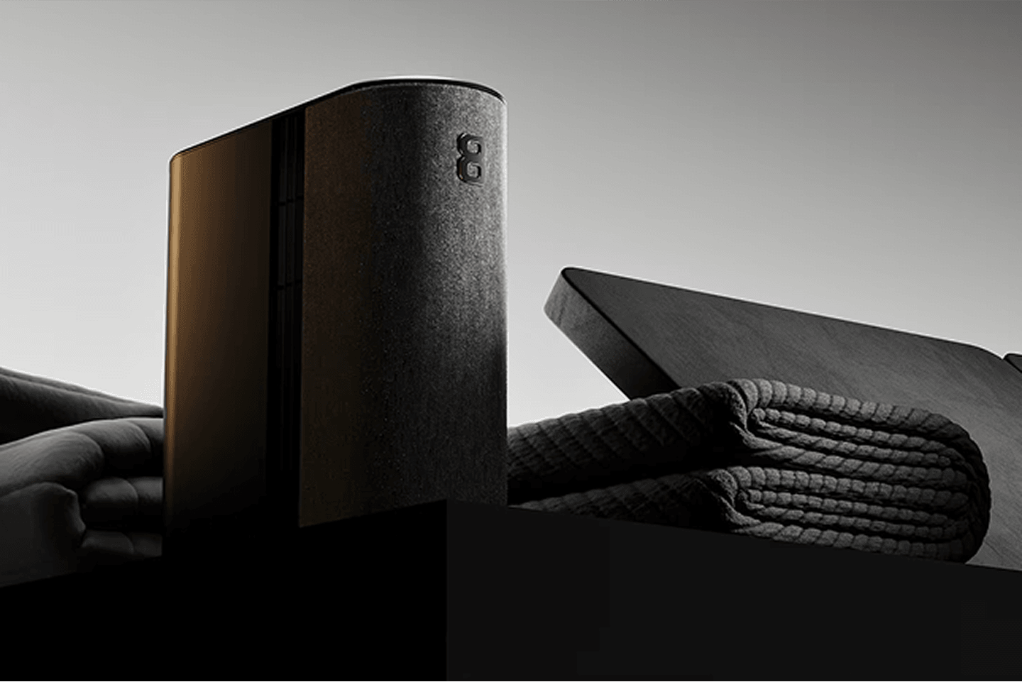 A sleek, modern speaker with a textured fabric finish sits on a table beside a folded, textured blanket and a closed laptop.