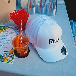White cap with "Rho" text next to a cocktail with an orange slice and striped straw on a table with cocktail menu and red stirrers.