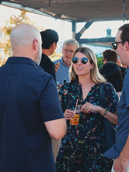 A group of people socializing outdoors, with a woman holding a drink and wearing sunglasses, engaged in conversation with two men.